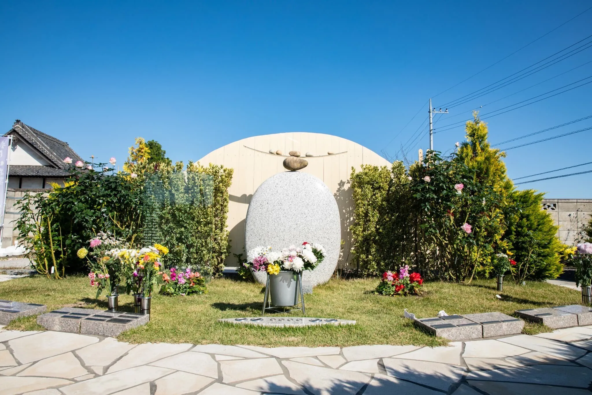 白い石碑と緑の植栽に囲まれた集合墓。明るい青空の下で花が供えられている。
